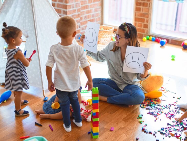 Beautiful psychologist and group of toddlers make therapy using emotions emojis around lots of toys at kindergarten