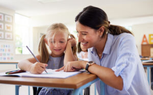 Teacher, learning and helping girl in classroom for drawing, studying or assessment. Teaching, development and kid or student with happy woman for education assistance with notebook in kindergarten.